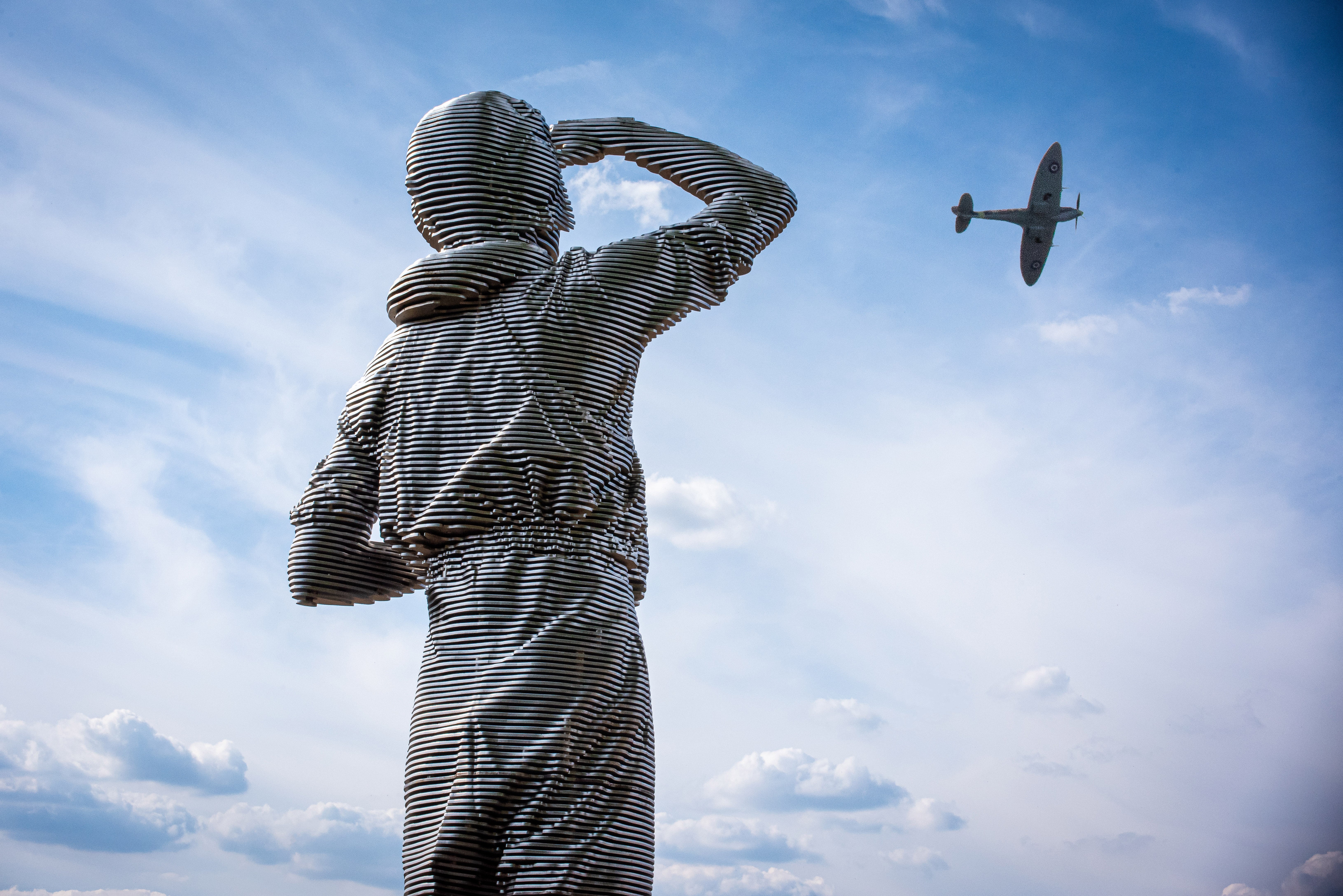 Image shows metal sculpture looking towards the sky with Spitfire flying overhead.
